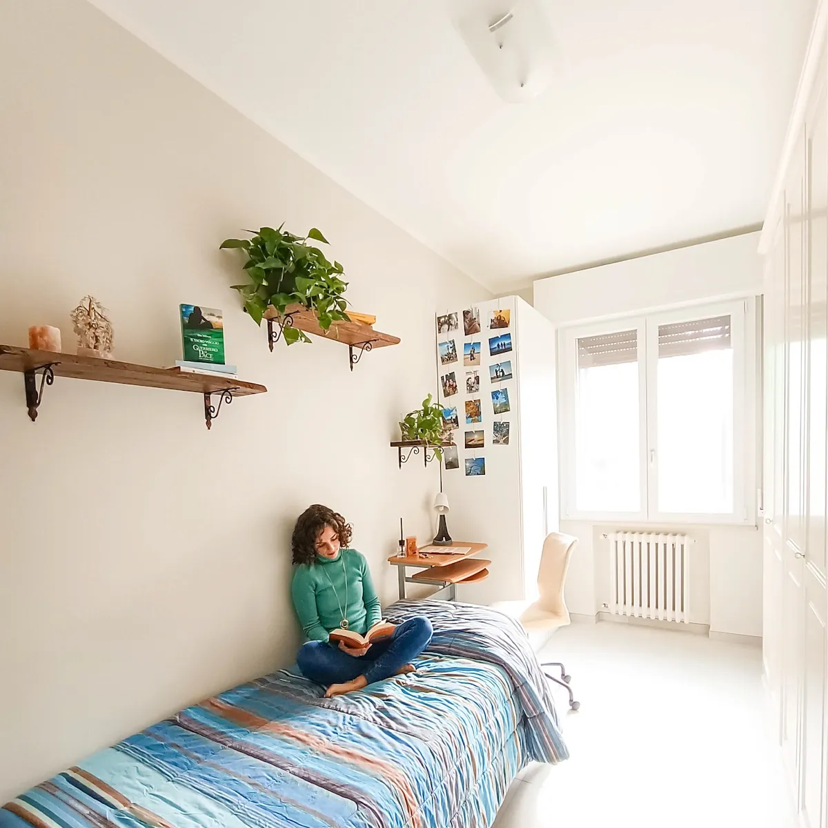 La stessa camera piccola dopo l'intervento, con mensole in legno, piante, parete di fotografie personali e donna che legge sul letto.