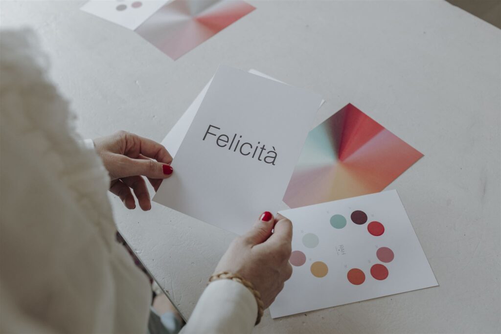 Mani con smalto rosso tengono le carte colore del test RAH su un tavolo bianco; una riporta la parola «Felicità»
