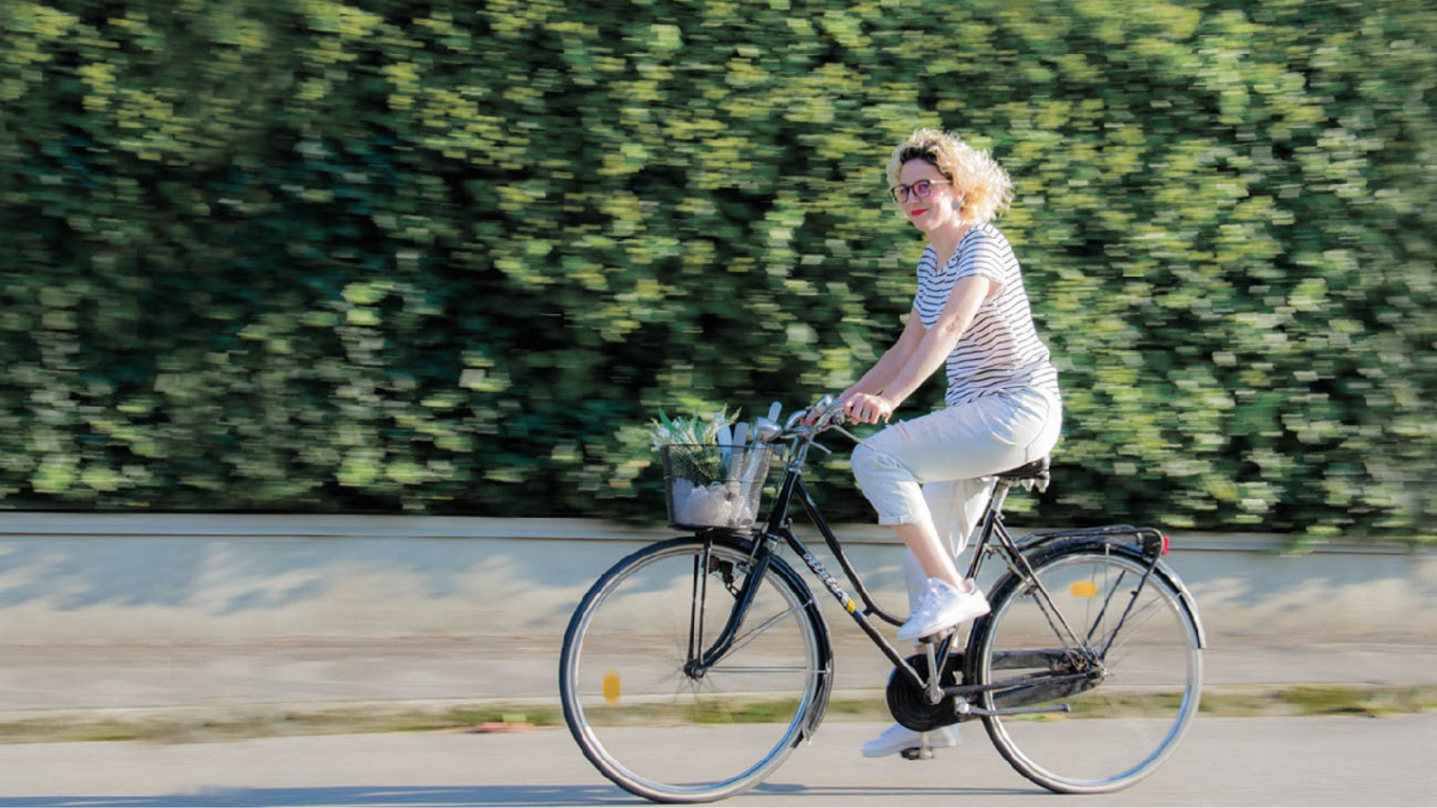 Francesca Campese, interior coach, pedala in bicicletta con un cesto di piante lungo una strada immersa nel verde.