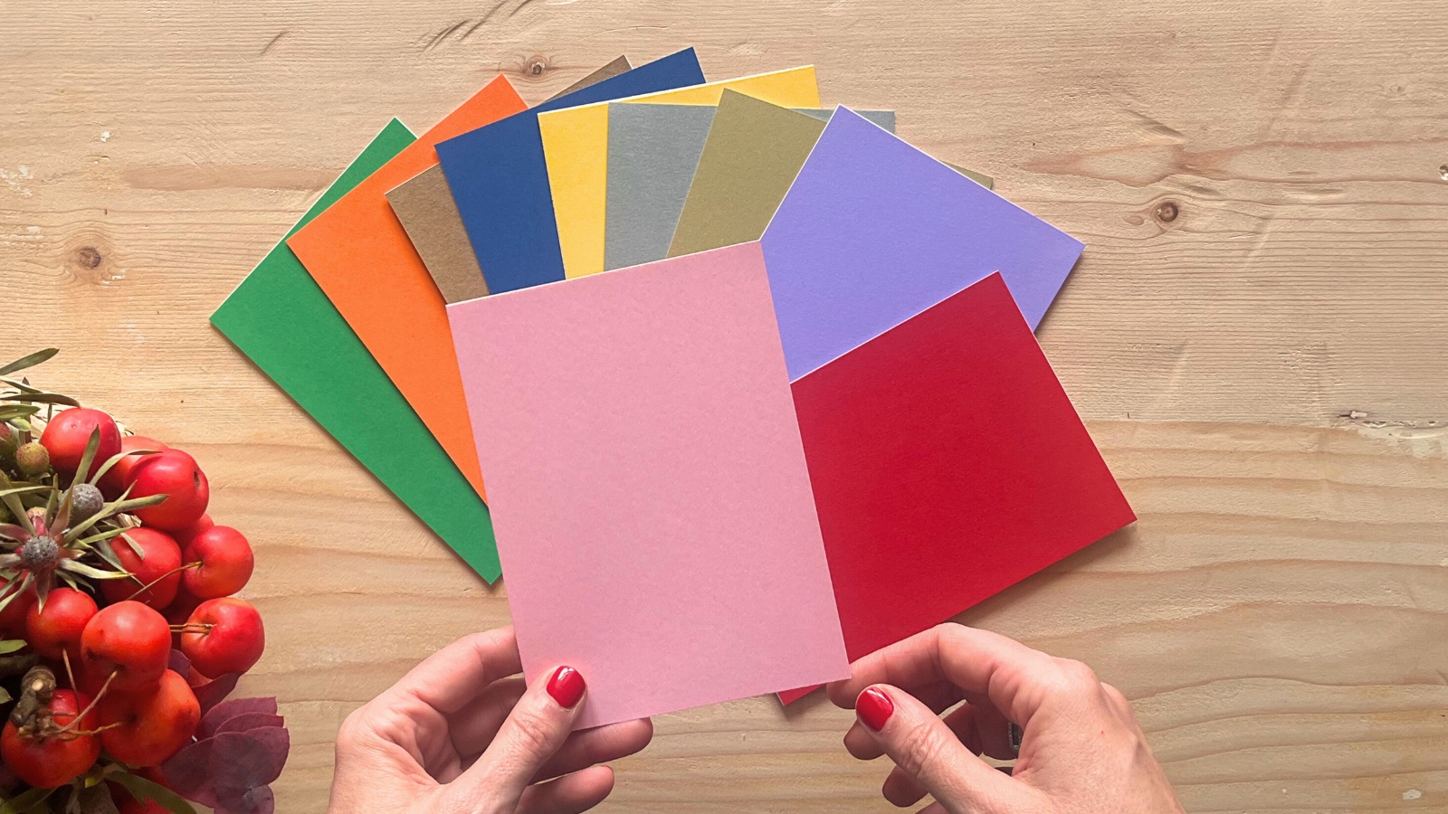 Le mani di una donna reggono un ventaglio di cartoncini colorati sopra un tavolo di legno, accanto a bacche rosse autunnali.